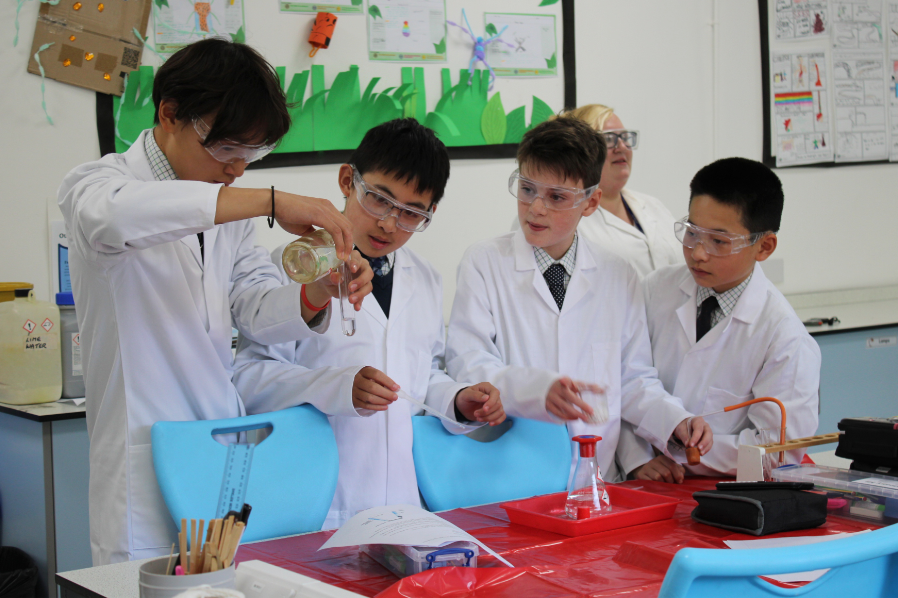 Four Horris Hill students are dressed in lab coats and goggles. They watch a member of the group carrying out a chemistry experiment.