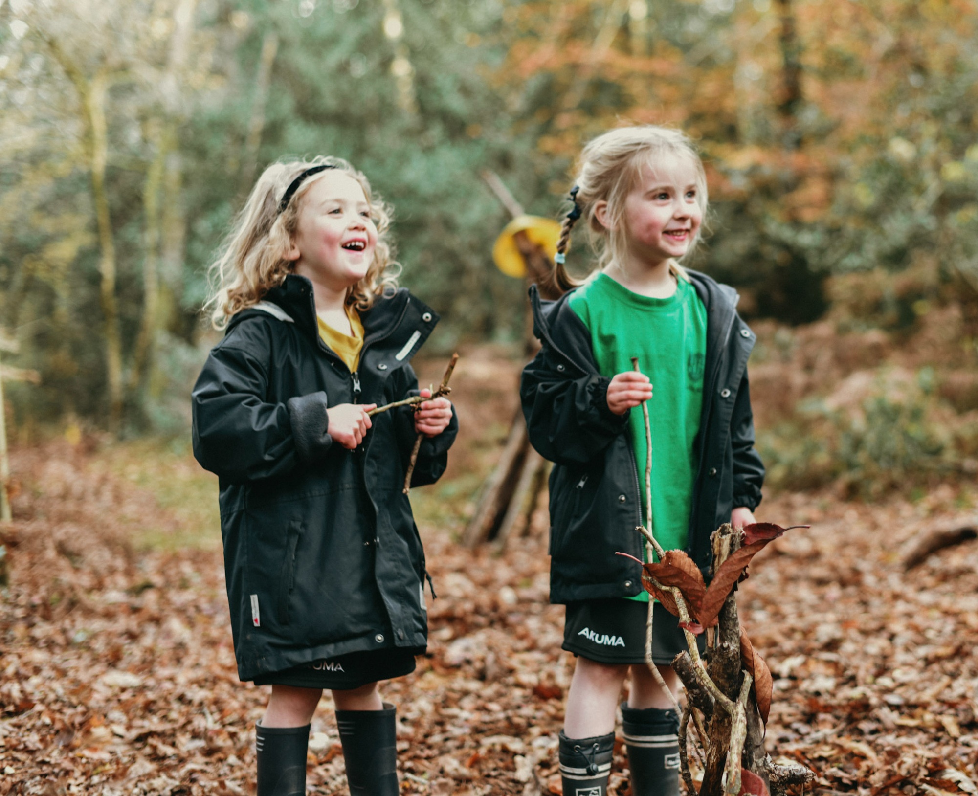 Two girls in Horris Hill School sports kit and raincoats stand in the woods. They are holding twigs and leaves and smiling.